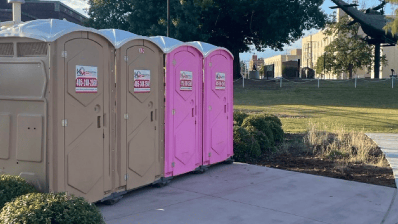 Four K&M porta potties lined up on a paved area beside a lawn